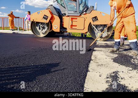 Workers are using rakes to level, set up layer of fresh tarmac to right measures, steamroller is flatting fresh asphalt Stock Photo