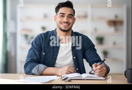 Cheerful arab guy in casual sitting at workdesk, taking notes Stock Photo