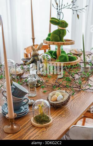 Stylish Easter table decor with candles, greenery, bunny figurines and natural elements in bright spring room interior. Cozy festive dining setup with Stock Photo