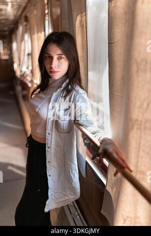 Young Woman StandinA young woman standing inside a train car, leaning on a handrail and looking at the camera. Warm sunlight enters through the window Stock Photo