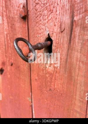 Close-up of a hand-forged key in a rustic keyhole on a red painted wooden barn door, Scandinavian rural detail. Stock Photo