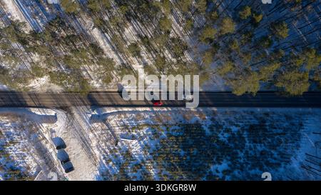 Aerial view of a two lane road in Poland through pine forest, three cars move on asphalt, two cars park on snowy pull off, long tree shadows cross sno Stock Photo