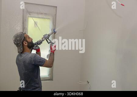 Construction worker applies a white coat, putty on the wall, using nozzle, plastering and skim coating walls spraying mixture under pressure Stock Photo