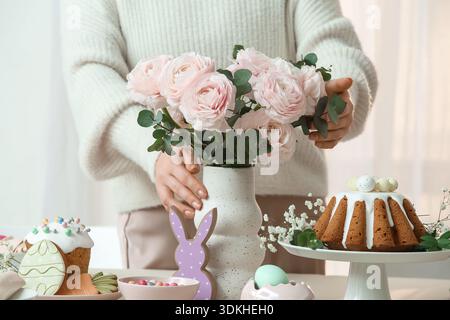 Woman holding vase with roses on table setting for Easter celebration in room. Closeup Stock Photo