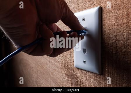 A close-up, high-detail shot of a hand plugging a blue RJ45 Ethernet cable into a white wall-mounted network port. Stock Photo