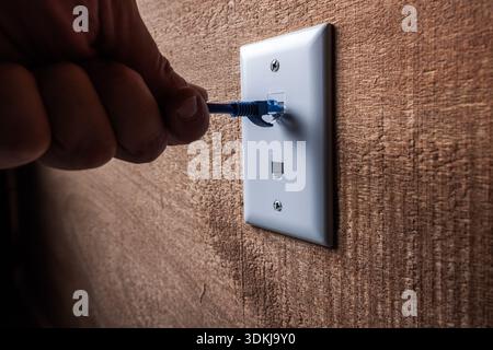 A close-up, high-detail shot of a hand plugging a blue RJ45 Ethernet cable into a white wall-mounted network port. Stock Photo
