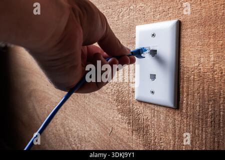 A close-up, high-detail shot of a hand plugging a blue RJ45 Ethernet cable into a white wall-mounted network port. Stock Photo