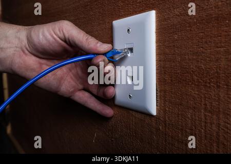 A close-up, high-detail shot of a hand plugging a blue RJ45 Ethernet cable into a white wall-mounted network port. Stock Photo