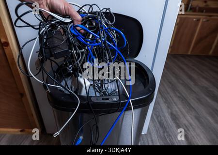 A high-angle conceptual shot of a hand discarding a large, tangled cluster of various electronic wires and cables into a stainless steel trash can. Stock Photo