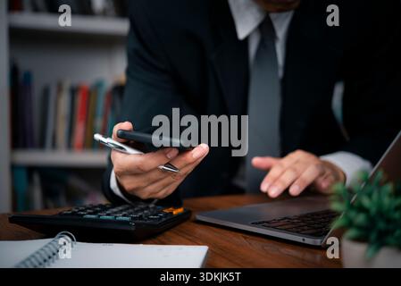 Businessman using smartphone and laptop at office desk with calculator, analyzing financial data, multitasking for corporate success and professional Stock Photo