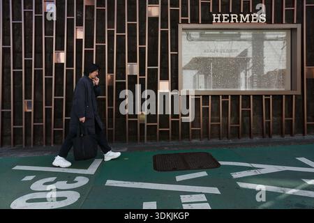 A man, using a cellphone, walks past the wall and window display of an Hermes Store in Omote-sando, Shibuya, Tokyo, Japan. Stock Photo