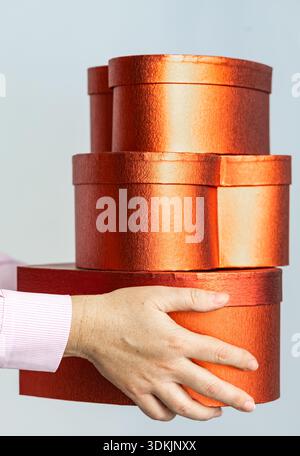 Close-up of a hand in pink shirt sleeve balancing stacked metallic red-orange round gift boxes, studio shot with copy space. Stock Photo