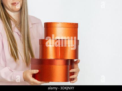 Blonde woman in pink shirt holds stacked metallic orange cylindrical gift boxes, studio shot with copy space on right. Stock Photo
