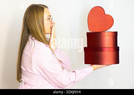 Horizontal wide angle eye level shot of a woman in profile expressing amazement while holding three red heart-shaped gift boxes on a white background Stock Photo