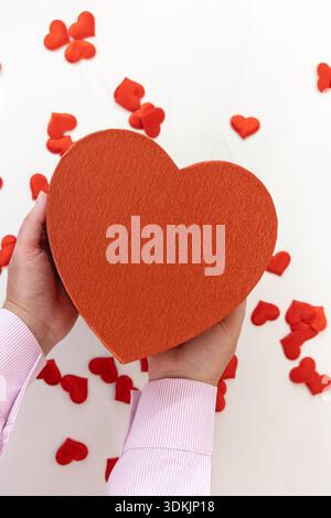 Vertical wide angle eye level shot of hands in a pink shirt presenting a large textured heart box surrounded by small red hearts on a bright surface Stock Photo