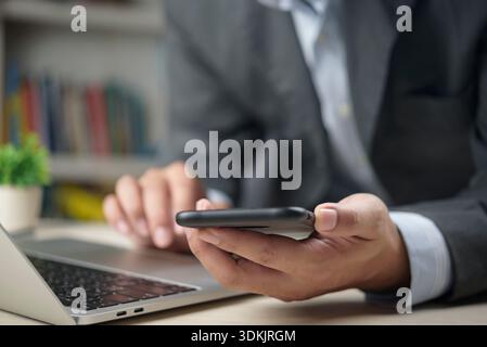 Man using mobile phone and laptop simultaneously in office. Business professional browsing internet, using social media for marketing, or checking fin Stock Photo