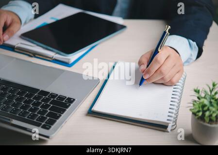 Business professional writes notes on notebook at office desk with laptop tablet paperwork for planning meeting agenda project management productivity Stock Photo