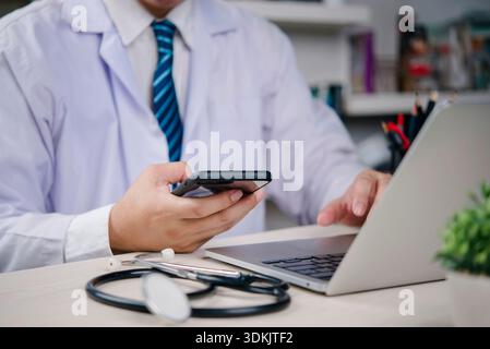 Healthcare medical Doctor with stethoscope works on laptop while checking smartphone to manage EHR records, telehealth messages, and patient data secu Stock Photo