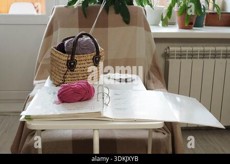 Home craft corner. Basket of yarn, open crochet tutorial book, and finished knitted flowers on white table against cozy chair. Stock Photo