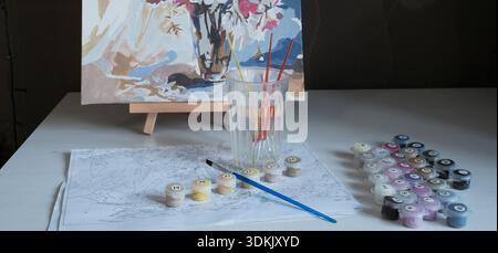 Creative workspace. Numbered painting scheme, set of acrylic colors, brushes in glass of water, and partially finished floral painting on small wooden Stock Photo