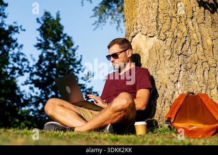 Man working on laptop outdoors while sitting under tree in casual outfit Stock Photo