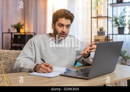 Young Indian man sitting at table joins online class through laptop, attentively listening to lecture, taking notes in notebook. Hispanic guy at home, focused on own task or learning activity nearby. Stock Photo