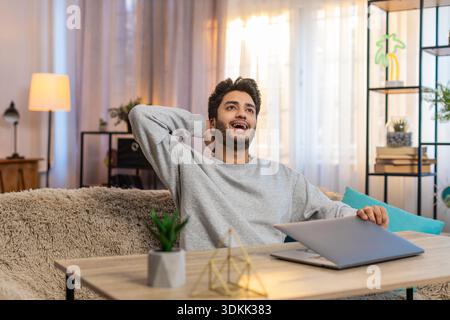Young Indian man sitting at table with laptop, smiling after completing successful project, closing laptop, and celebrating. Hispanic Hindu guy relaxing taking a break on sofa in living room at home. Stock Photo