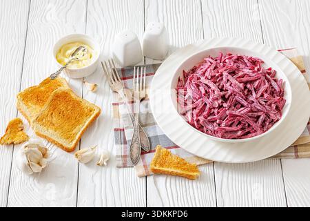 Traditional Eastern European beetroot salad with garlic and mayonnaise in a white bowl on a light wooden table with toasted bread and forks, horizonta Stock Photo