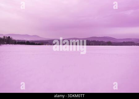 A wide view of a snow-covered field with distant mountains and houses in purple tones, overcast sky Stock Photo