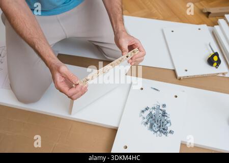 man carefully aligns two furniture panels with dowels on a wooden floor surrounded by tools, hardware, and assembly instructions in a bright room Stock Photo