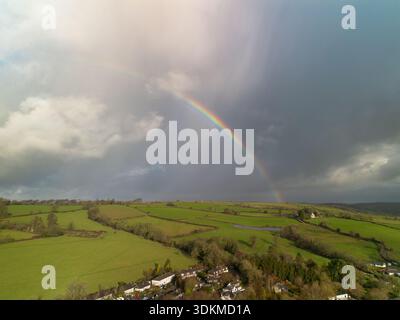 Bright rainbow over green countryside fields and a rural village under dramatic storm clouds. Stock Photo