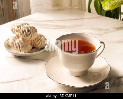 Tea with Zephyr Marshmallows. Cozy tea time with a cup of hot black tea and soft airy Russian zephyr marshmallows on a light table near the window. Stock Photo