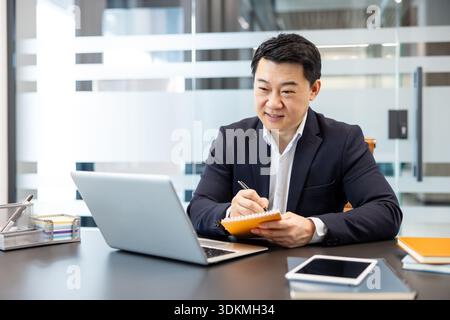 Businessman working in an office, focusing on his laptop screen while actively writing notes in a colorful notebook, demonstrating professional dedication and multitasking during a virtual meeting Stock Photo