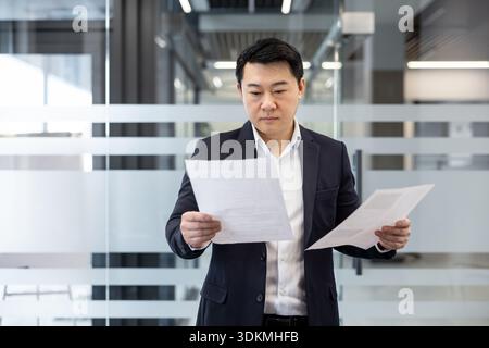 Asian businessman standing in a contemporary office setting, meticulously reviewing important paperwork and corporate reports, focusing on contracts and financial data Stock Photo