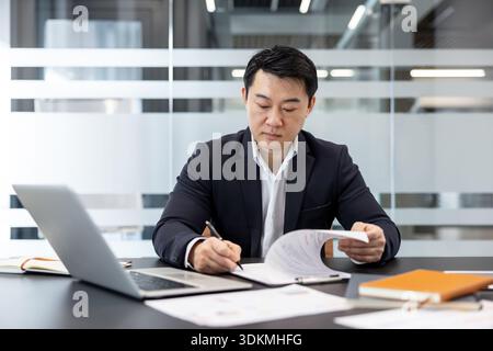 Asian businessman in a modern office reviewing financial reports, signing contracts and working on a laptop with focused concentration on management and accounting tasks Stock Photo