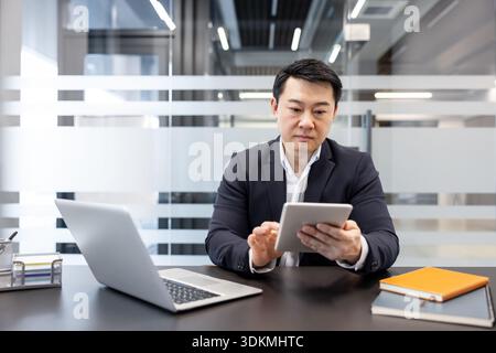 Asian businessman working in an executive office, focusing on his digital tablet while a laptop sits open on the dark desk, symbolizing productivity and modern business communication Stock Photo