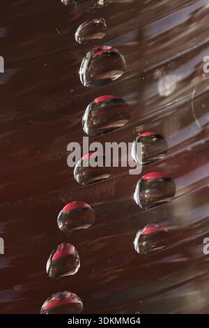 Abstract macro of clear water droplets on a dark, ribbed surface with vibrant red reflections. Artistic close-up of texture, symmetry, and moisture. Stock Photo