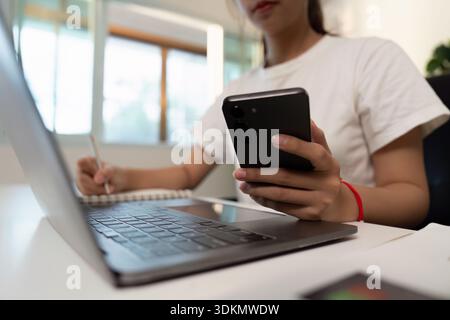 Multitasking. Young individual using smartphone while taking notes on laptop. Stock Photo