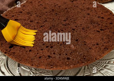 Applying coffee syrup on to a half of a homemade gluten free sponge cake with a cooking silicone brush, soft focus Stock Photo