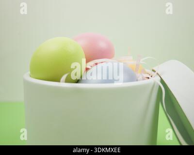 Colorful Easter eggs in shades of green, pink, and blue are nestled in a white container filled with decorative straw, set against a soft green backgr Stock Photo