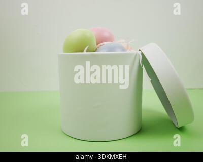 White round box partially open reveals pastel colored Easter eggs in shades of green, pink, and blue, placed on a green surface with a light green bac Stock Photo
