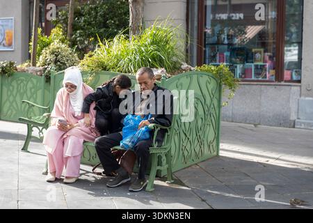 Moscow, Russia, July 17, 2025 Three people Muslim women in pink hijab and elderly man sitting on ornate green metal bench in Moscow, smartphone browsi Stock Photo