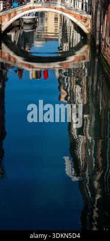 Venice, Italy. February 1, 2026. The reflection of an arched bridge and historic buildings appears in calm canal water in Venice during Carnival Stock Photo
