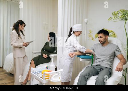 Wide shot of medical professional in uniform and gloves prepares male patient arm for blood draw while colleague having consultation with client in Stock Photo