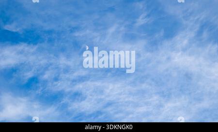 heaven skies blue and clear, with thin, puffy, transparent clouds. and moon is visible through them. sun shines brightly, making sky even brighter. sc Stock Photo