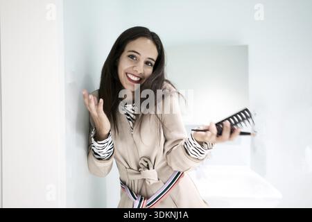 Carefree amused vibes of woman in a beige blazer playfully holding makeup and hairbrush while laughing in the bathroom, having applied makeup, applyin Stock Photo