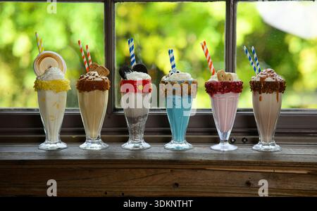 A row of six milkshakes with different toppings and colors. The milkshakes are lined up on a wooden counter Stock Photo