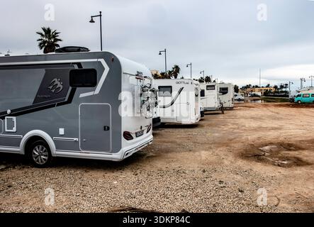 A line of modern motorhomes and camper vans are parked in a gravel lot near the coast under a cloudy sky in Valencia, Spain. Stock Photo