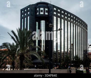 The modern Edificio Europa in Valencia features a dark, reflective glass facade and distinctive curved architectural lines near a palm-lined street. Stock Photo