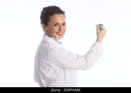 A professional female physician in a white lab coat smiles while holding a marker, appearing to explain medical concepts or write on a transparent boa Stock Photo